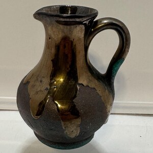 May include: A small, decorative ceramic pitcher with a dark brown base and a metallic gold and bronze glaze. The pitcher has a handle and a spout. The overall shape is rounded, with a slightly flared opening. The pitcher is displayed on a clear shelf.