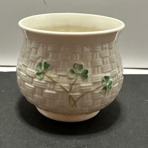 May include: A small, off-white ceramic pot with a basket weave design. The pot features three green shamrock designs. The pot has a rounded base and a wide opening. The pot is likely for decorative purposes.