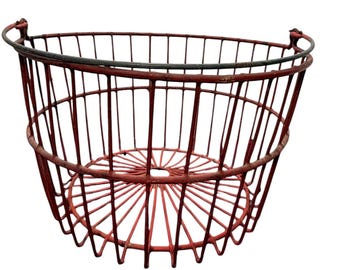 May include: A round, red wire basket featuring a sturdy metal frame with vertical slats and a circular base. The basket includes a dark, weathered metal rim at the top and two small, integrated metal loops on opposite sides for handling.
