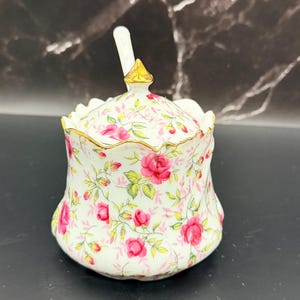 May include: A white ceramic sugar bowl with a lid and spoon, adorned with a pink rose floral pattern. The rim and handle are accented with gold trim. The spoon rests on top of the lid.