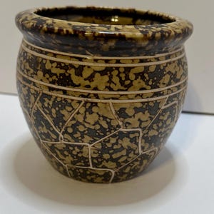 May include: A small, round ceramic pot with a speckled brown and tan pattern. The rim and top edge are a darker brown. The pot has a textured, mosaic-like design. The pot is likely used for decorative purposes.