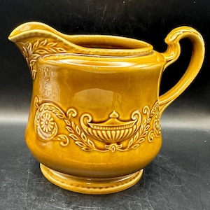 May include: A brown ceramic pitcher with a decorative floral design. The pitcher has a wide base and a rounded body with a spout and a handle.