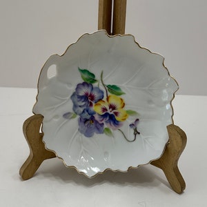 May include: A white porcelain dish shaped like a leaf with a gold trim. The dish features a hand-painted design of purple and yellow pansies.