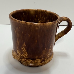 May include: A brown ceramic mug with a handle. The mug has a speckled, glazed finish with shades of brown and tan. The mug is cylindrical with a slightly flared rim and a rounded base.