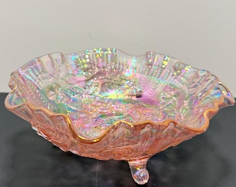 May include: A peach-colored iridescent glass bowl with a ruffled, scalloped edge and a textured floral pattern. The bowl is supported by three small, curved feet and features a reflective, multi-colored sheen across its surface.