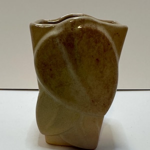 May include: A tan ceramic vase with a unique, twisted design. The vase has a rectangular base and a wavy top, with a light brown color gradient. The vase is a decorative item, suitable for displaying flowers or as a standalone art piece.