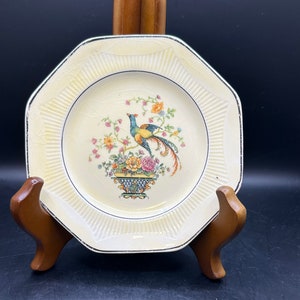 May include: A white octagonal plate with a gold rim and a floral design featuring a green, blue, and yellow bird in a basket.