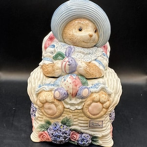 May include: A ceramic cookie jar shaped like a bear wearing a blue and white hat and dress, sitting in a wicker chair with flowers and a "Hand Painted" label.