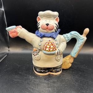 May include: A ceramic teapot shaped like a cat wearing a chef's hat and holding a cake. The teapot is decorated with blue, red, and yellow details.