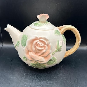 May include: A white ceramic teapot with a rose design. The teapot has a brown handle and a lid with a rose bud on top. The teapot is decorated with pink and green roses.