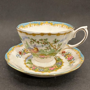May include: A white teacup and saucer set with a detailed bird and floral design. The cup and saucer have a blue and gold trim. The teacup has a curved handle. The set is made of porcelain.