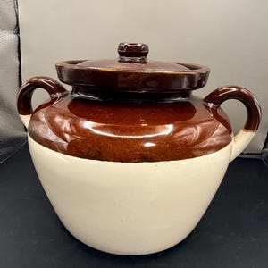 May include: A two-toned ceramic crock pot with a lid. The top half is a glossy brown, and the bottom half is a creamy white. It has two curved handles and a small knob on the lid.