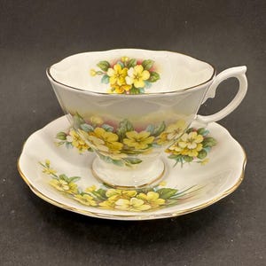 May include: A white teacup and saucer set with a floral design. The teacup and saucer are decorated with yellow flowers and green leaves. The rims are trimmed with gold. The teacup has a handle.