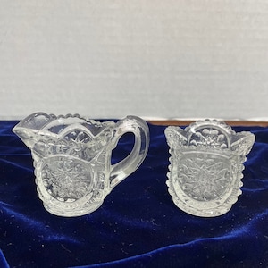 May include: A set of two clear glass creamer and sugar bowls with a cut glass design. The bowls are on a blue velvet surface.