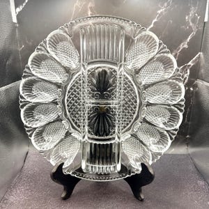 May include: A clear, vintage glass serving dish with a central rectangular section and a circular outer rim with scalloped edges. The dish is supported by a black stand, showcasing its intricate design and elegant form. The dish is designed to hold appetizers.