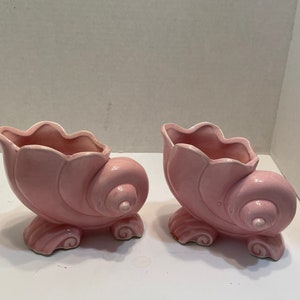 May include: Two pink ceramic seashell-shaped planters with white accents. The planters are in the shape of a conch shell with a curled lip and a detailed spiral pattern.