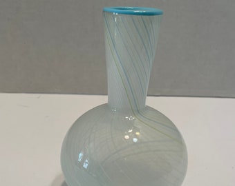May include: A small, white glass vase with a blue rim and swirling white and green lines. The vase has a round base and a narrow neck.