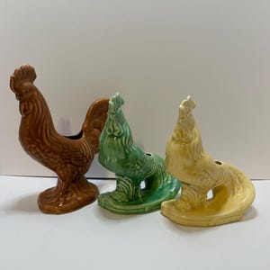 May include: Three ceramic rooster figurines in shades of brown, green, and yellow. The roosters have detailed feather textures and are standing upright. The figurines are decorative and likely for home decor.