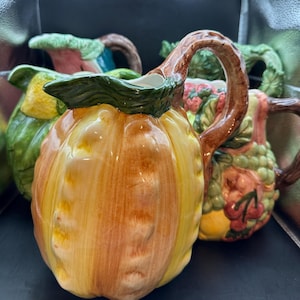 May include: A set of four ceramic pitcher-style serving pieces, each decorated with a different fruit or vegetable design. The pitchers are in shades of green, yellow, orange, and brown.