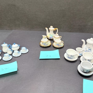 May include: Three sets of miniature porcelain tea services featuring white ceramic with blue floral patterns and gold trim. Each set includes a teapot, creamer, sugar bowl, and multiple teacups with matching saucers, arranged with blue paper labels.