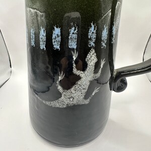 May include: A black and green ceramic pitcher with a white lizard design and a white and blue floral pattern around the middle.