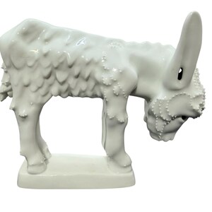 May include: A white ceramic figurine of a donkey standing on a rectangular base. The donkey has a glossy finish with textured, raised details along its back, legs, and head, featuring long ears and a downward-facing head position.