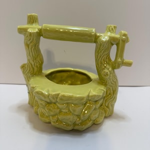 May include: A ceramic well-shaped planter in a light green color. The planter has a stone-like texture around the base and a wooden-style structure above with a rolling handle. The well is designed to hold small plants or flowers.