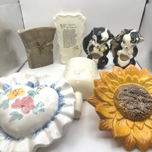 May include: A collection of seven ceramic wall decor pieces. The pieces include a heart-shaped pillow with floral designs, a sunflower, a white vase with a floral design, two black and white skunk planters, and a rectangular white plaque with a poem.