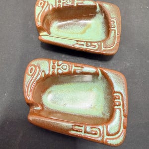 May include: Two rectangular ceramic trays featuring a glossy brown outer border and a recessed, light green glazed center. The brown edges are decorated with raised, embossed geometric patterns that wrap around the sides of each tray.