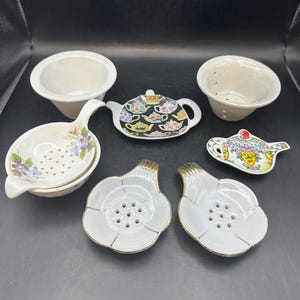 May include: A set of seven white porcelain tea infusers, including a teapot, a flower-shaped infuser, and a round infuser with a handle. The infusers are decorated with floral patterns and gold accents.