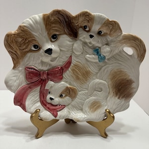 May include: A ceramic serving dish shaped like three dogs. The largest dog is wearing a pink bow and the two smaller dogs are peeking out from behind the larger dog. The dish is on a gold stand.
