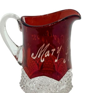 May include: A ruby red glass creamer pitcher featuring a clear glass handle and a clear base with a textured diamond pattern. The name Mary is etched in cursive script on the front of the smooth red upper section of the pitcher.