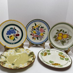 May include: Four ceramic plates with different fruit designs. The plates have a white background with colorful fruit designs, including grapes, cherries, peaches, and flowers. The plates have a yellow, green, and blue border.