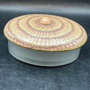 May include: An oval-shaped trinket box with a white base and a lid with a pink, brown, and white patterned design. The lid has a gold trim and a small knob on top.