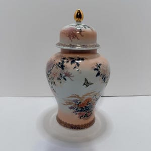 May include: A decorative ceramic ginger jar with a lid. The jar features a peach and white background with floral and butterfly designs. The lid is topped with a gold finial and has a decorative border.