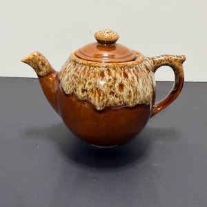 May include: A brown ceramic teapot with a lid and handle. The teapot has a rounded body with a lighter brown, textured glaze around the top. The spout and handle are also brown. The lid has a small knob on top.