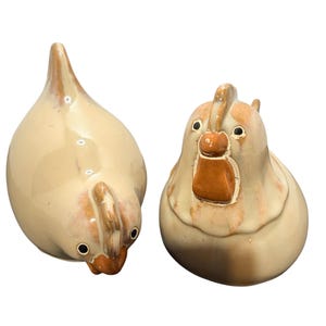 May include: Two ceramic chicken figurines with a glossy cream-colored glaze and brown accents on the combs, wattles, and beaks. One chicken is positioned looking downward, while the other is facing forward with a prominent comb and wattle detail.