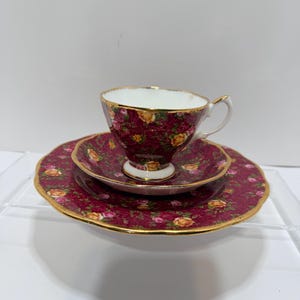 May include: A vintage tea set featuring a burgundy background with a floral pattern of yellow and pink roses. The set includes a teacup, saucer, and a dessert plate, all with gold trim. The teacup is perched atop the saucer.