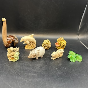 May include: A collection of small, detailed animal figurines. The set includes a giraffe, dolphin, frog, pig, and other creatures. The figurines are in various colors, including brown, beige, and green. They appear to be made of ceramic or a similar material.