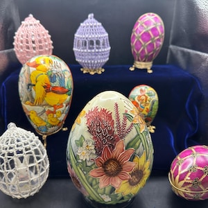 May include: A collection of eight decorated Easter eggs. The eggs are displayed on a blue velvet surface. Some of the eggs are painted with floral designs, while others are covered in intricate patterns. One egg features a painted image of a duck and chicks.