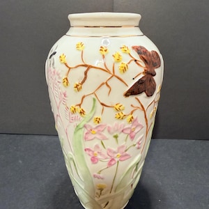 May include: A cream-colored ceramic vase featuring raised relief decorations of pink flowers, green leaves, and brown branches with yellow buds. A large brown butterfly is perched on the branches. The rim has a thin gold-colored metallic band.