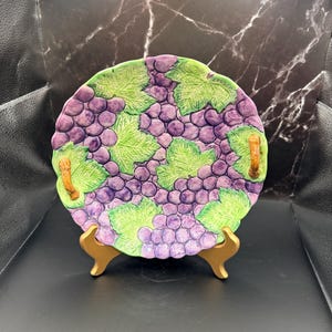 May include: A decorative ceramic plate featuring a grape design. The plate is round with a scalloped edge and two small handles. The grapes are purple, and the leaves are green. The plate is displayed on a gold-colored stand.