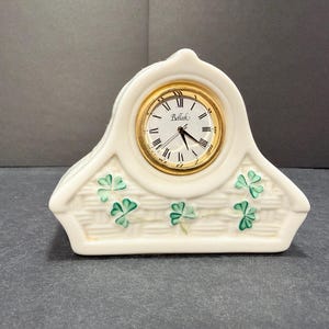 May include: A cream-colored porcelain mantel clock with a basket-weave texture and hand-painted green shamrock accents. The clock face features a gold-tone bezel, black Roman numerals, and the brand name Belleek printed in black script.