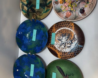 Könnte beinhalten: Eine Sammlung von sechs dekorativen Emaille-Tellern. Die Teller weisen verschiedene Designs auf, darunter Blumenmuster, abstrakte Formen und einen Roadrunner-Cartoon. Zu den Farben gehören Blau, Grün, Braun und Gold. Jeder Teller hat ein einzigartiges Design und Farbschema.