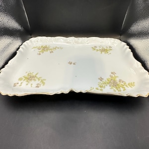 May include: A white porcelain serving tray with a scalloped edge and a gold trim. The tray is decorated with a delicate floral pattern in shades of green and purple.