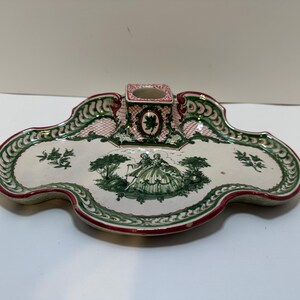 May include: A decorative ceramic tray with a central square holder. The tray features a scalloped edge with red trim and green and white floral patterns. A central scene depicts figures in a garden setting.