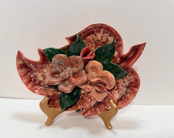 May include: A leaf-shaped ceramic dish featuring a variegated red and white glaze. The center is adorned with three-dimensional sculpted flowers in shades of pink and peach, surrounded by glossy dark green ceramic leaves.