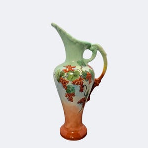 May include: A tall, vintage ceramic pitcher with a curved spout and handle. The pitcher features a light green top, transitioning to a peach and orange base. It is decorated with red grape clusters and green leaves.