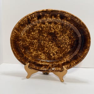 May include: An oval-shaped brown ceramic platter with a speckled pattern and decorative rim. The platter is displayed on a gold-colored stand, showcasing its rustic design. This vintage-style serving dish is suitable for various culinary presentations.