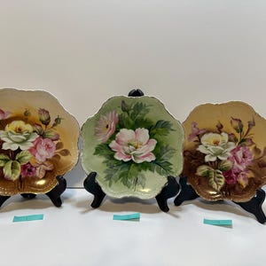 May include: Three decorative porcelain plates with scalloped edges, each featuring a floral design. The plates are supported by black stands. The flowers are painted in shades of pink, yellow, and white, set against a gold and green background.